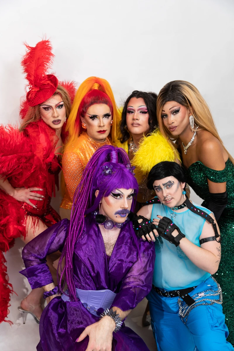 Group portrait of drag performers wearing colourful outfits and makeup against a studio backdrop.