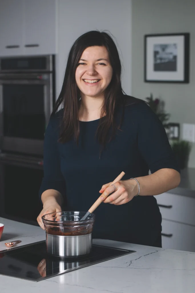 Woman smiles while stirring melted chocolate in a pot in a kitchen.