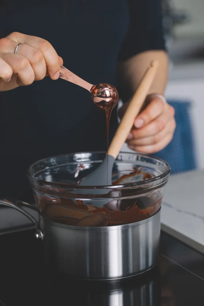 Spoon lifts melted chocolate above a pot while another hand stirs with a spatula.