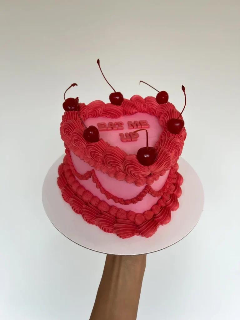 Heart-shaped pink cake topped with cherries and the words “Eat Me Up.”
