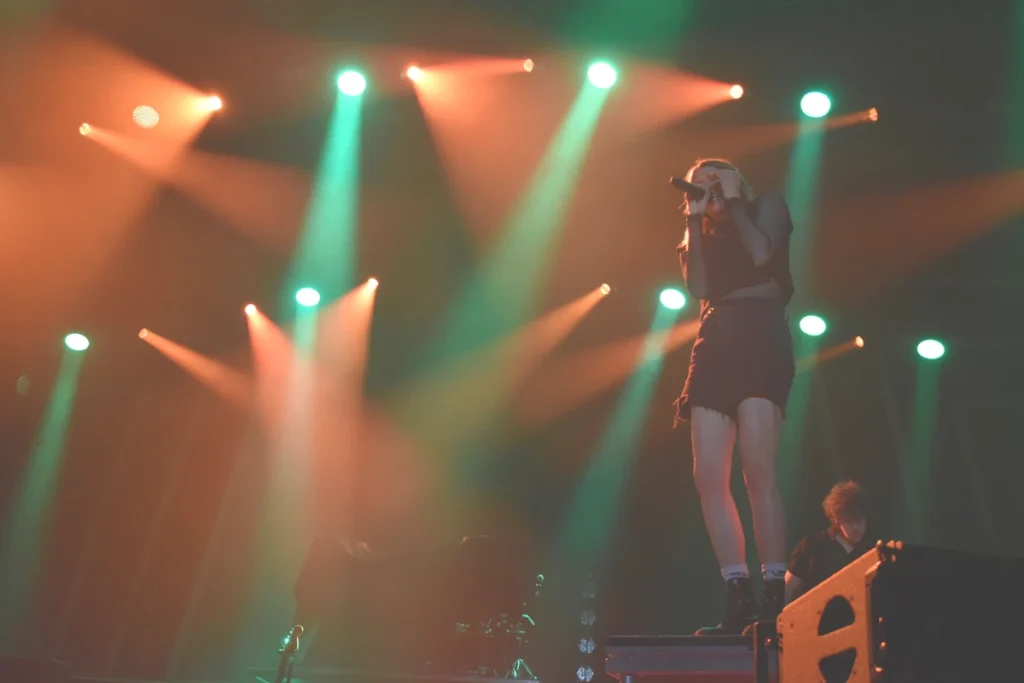 Singer stands on a platform performing into a microphone under colourful stage lights.