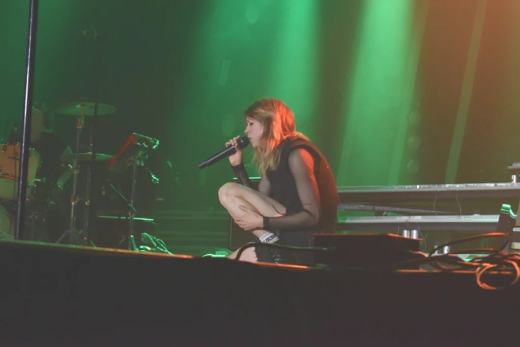 Singer crouches onstage while performing into a handheld microphone under green lighting.