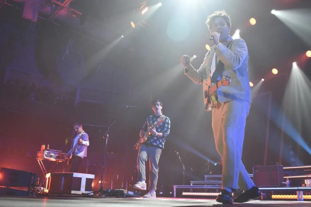 Band performs onstage with guitars and microphone under bright concert lighting.