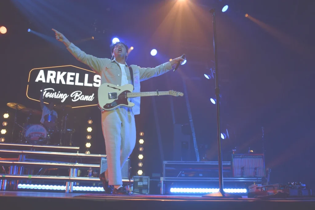 Arkells frontman stands facing the audience with arms raised while performing onstage.