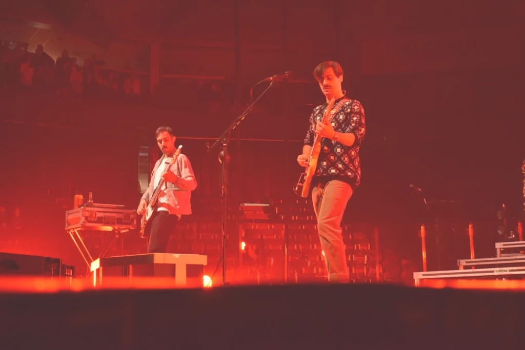 Two Arkells band members play guitar and bass onstage under red concert lighting.