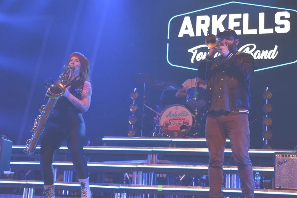 Saxophonist and trumpet player perform onstage with the “ARKELLS Touring Band” sign visible behind them.