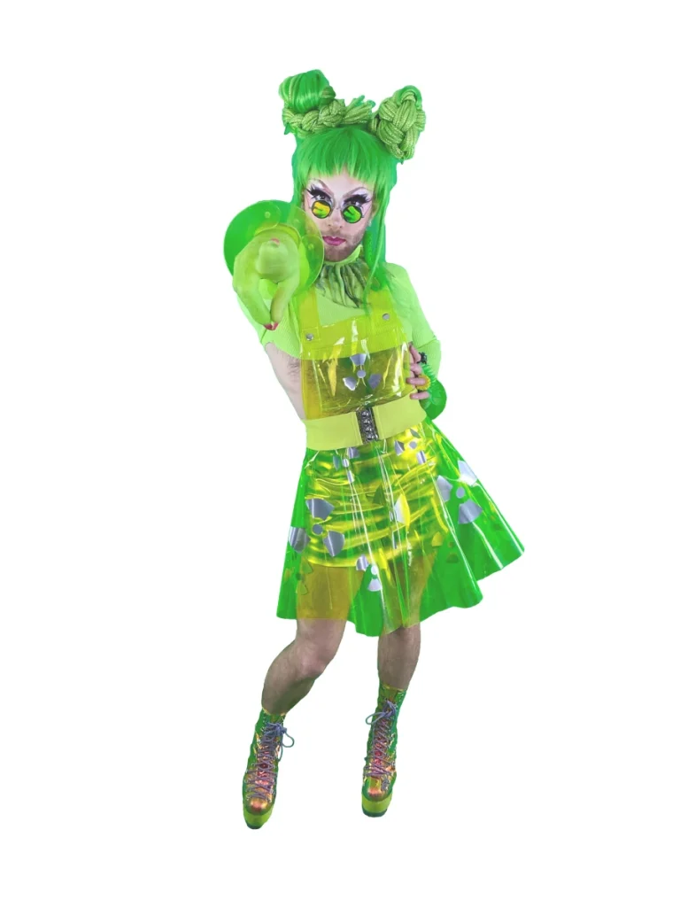 Drag performer wearing a neon green outfit, green wig, and reflective glasses posing against a white background.