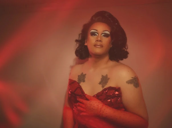 Drag performer in a red dress poses under warm red stage lighting.