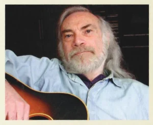 Portrait of Hank Davis with long grey hair and beard holding an acoustic guitar.