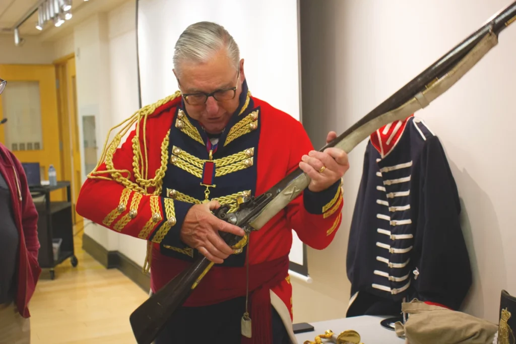 Man in historical military uniform demonstrates a long rifle during a presentation.