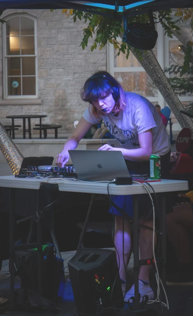 DJ performs at a laptop and mixer table during an outdoor set.