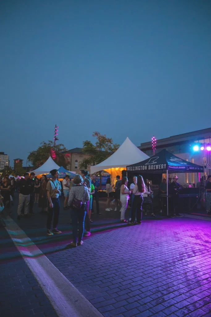 Festivalgoers gather among vendor tents and stage lighting at an outdoor Guelph Jazz Festival evening event.