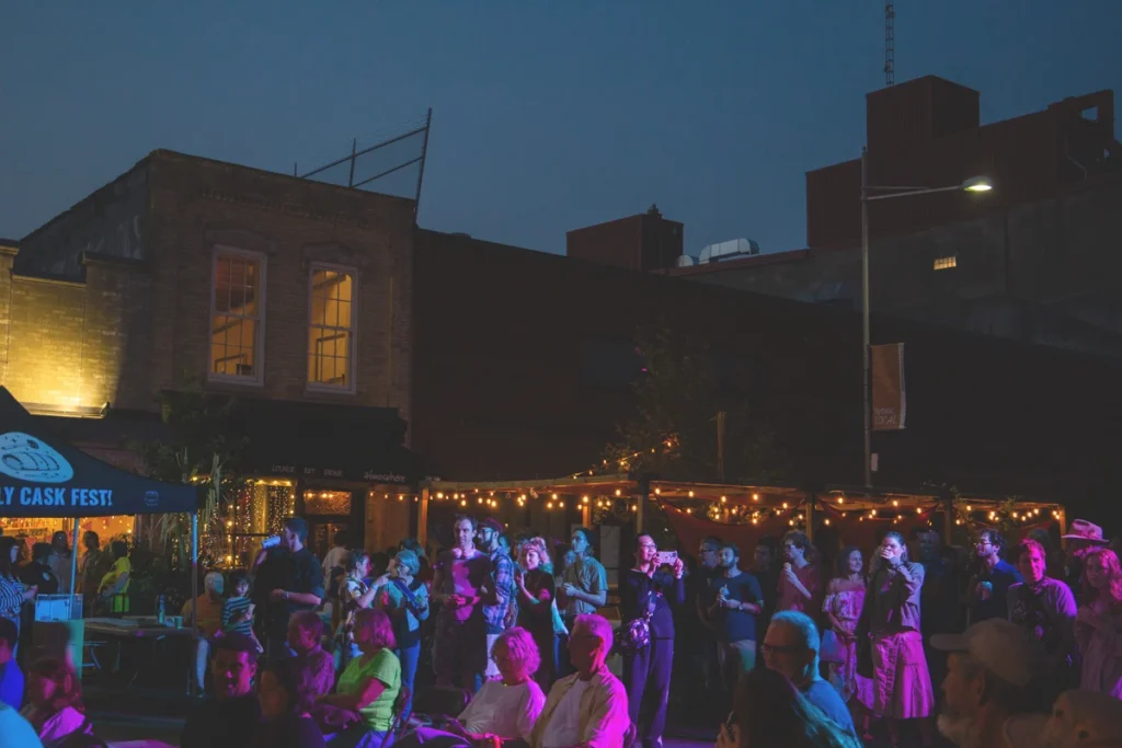 Audience members stand and sit in a courtyard watching a nighttime jazz performance under string lights.