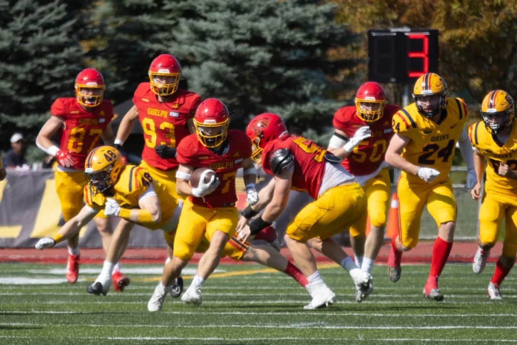 University of Guelph football player runs with the ball while Queen’s defenders close in during a game.