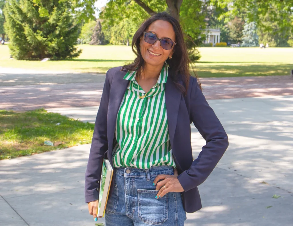 Madhur Anand wearing glasses and a green striped shirt stands outdoors holding a book, smiling at the camera.