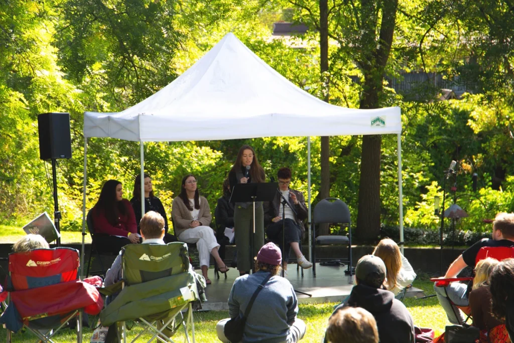Speaker stands at a podium under a white canopy while an audience sits on lawn chairs in a park setting.
