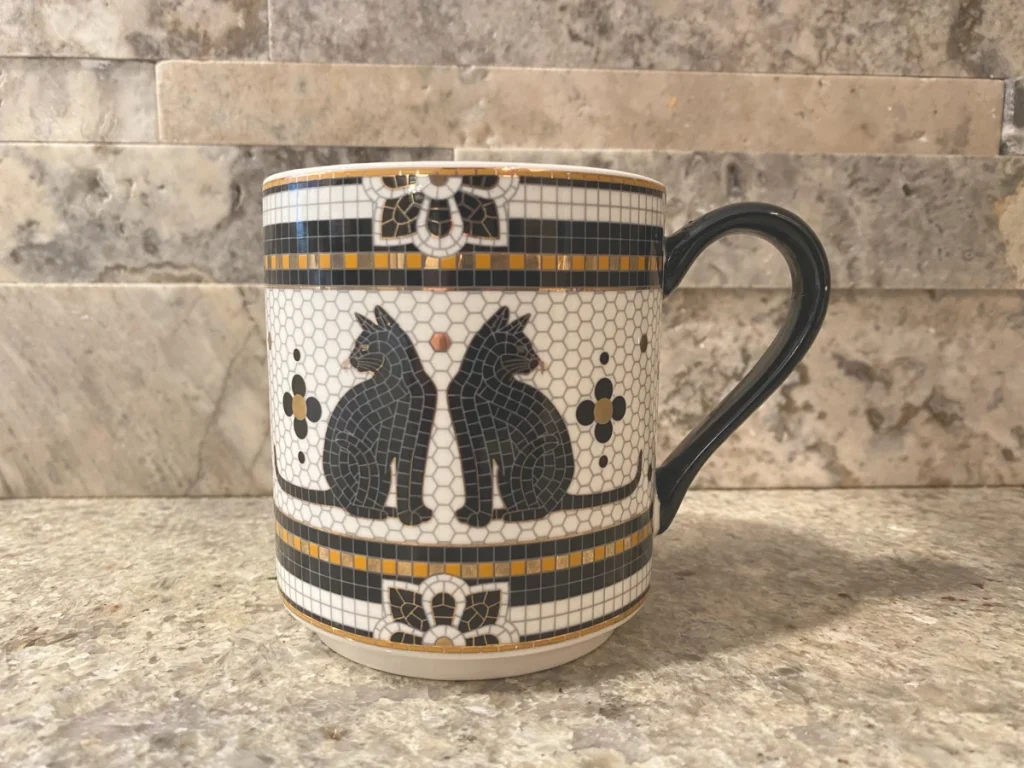 Ceramic mug with mosaic-style pattern and black cat illustrations on a tiled kitchen backsplash background.