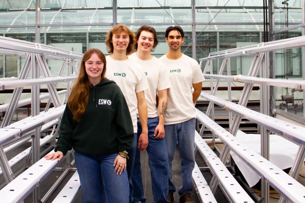 Four students wearing ESW clothing stand smiling between rows of metal hydroponic growing racks inside a greenhouse.