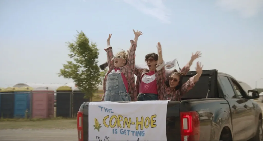 Three people wearing matching plaid shirts cheer with their arms raised while riding in the back of a pickup truck carrying a handmade sign.