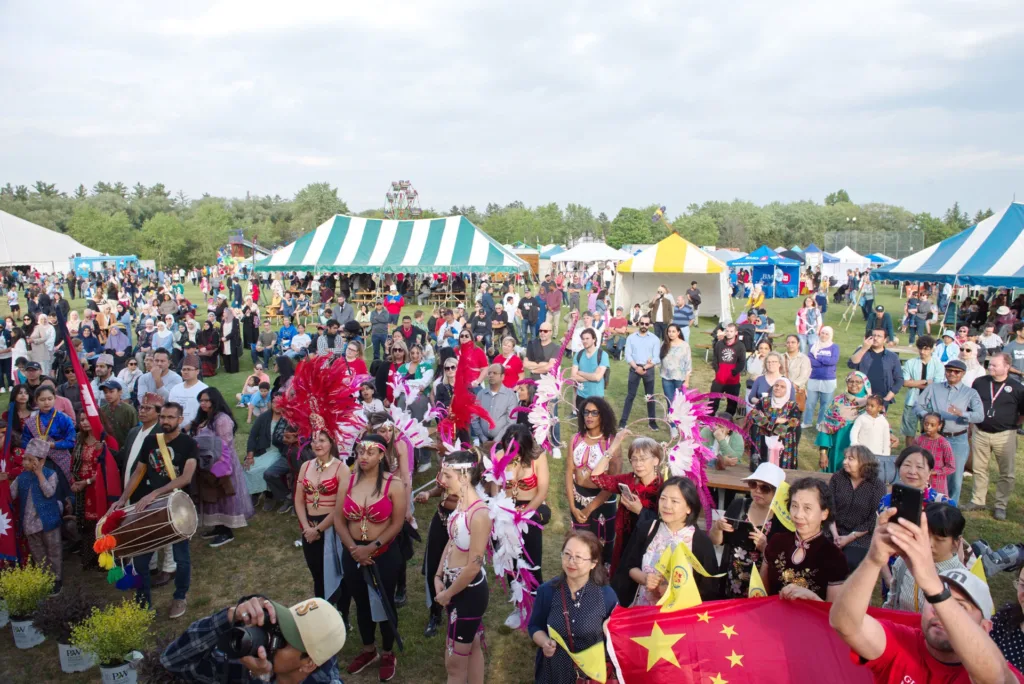 Festival attendees gather on a grassy field during the Guelph and District Multicultural Festival, with dancers in feathered costumes and people holding international flags near the front of the crowd.