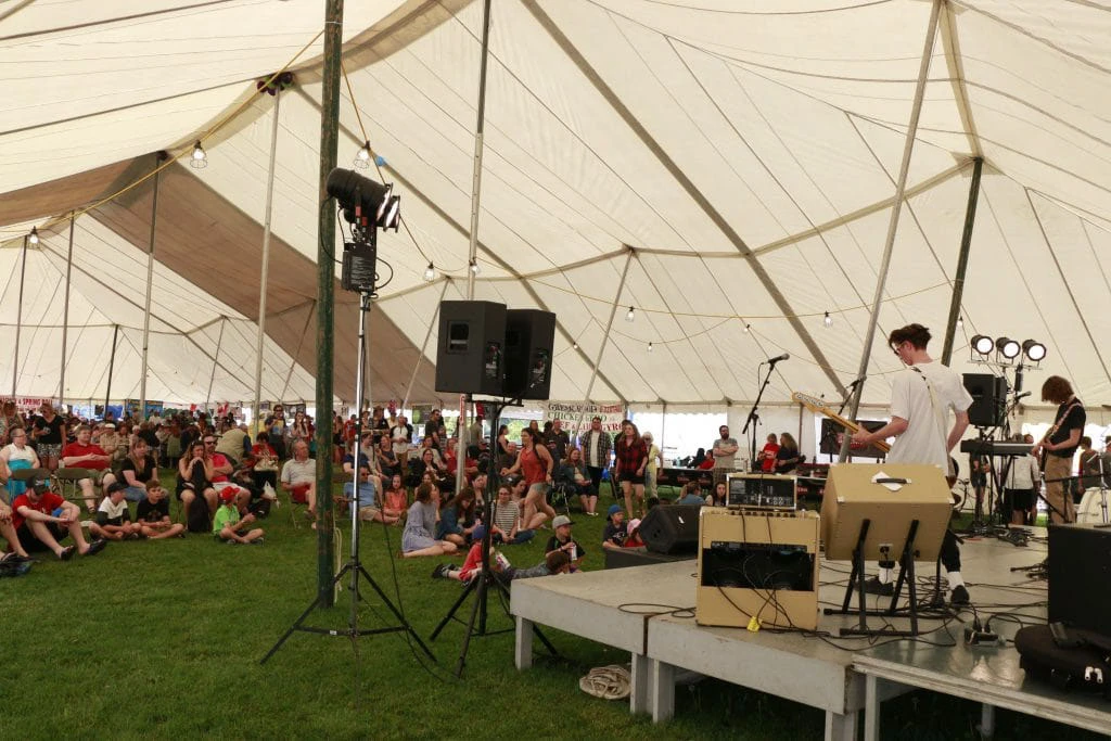 A band performs onstage inside a large festival tent while audience members watch from the grass and folding chairs.