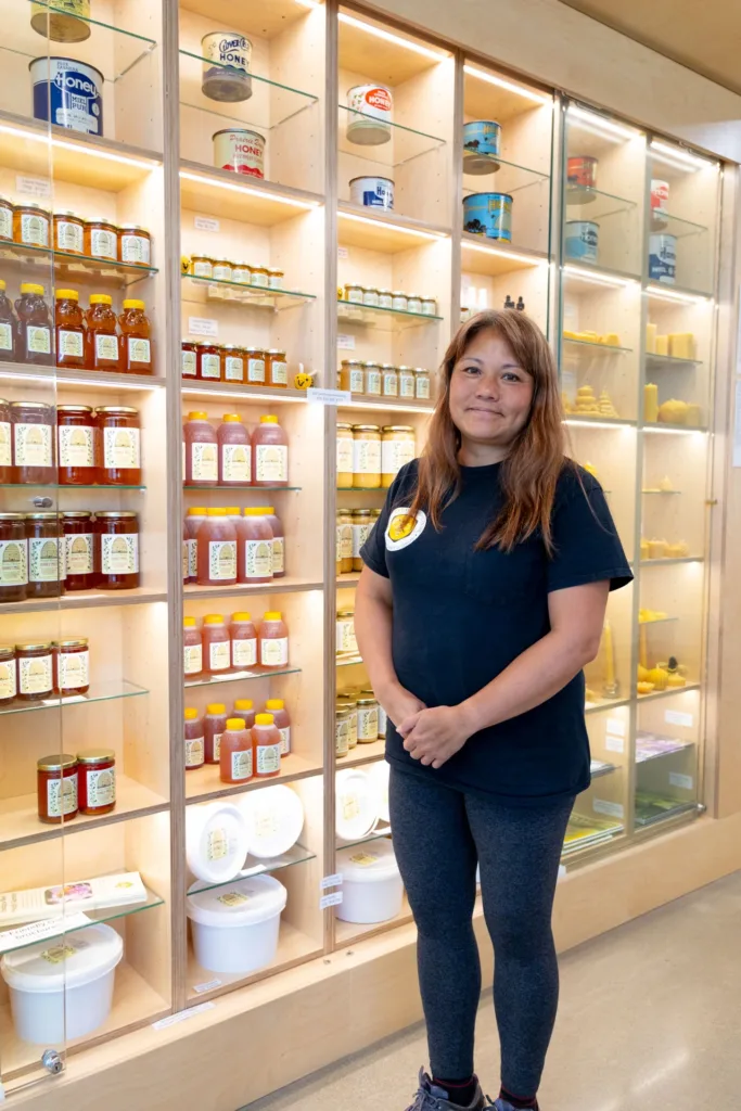A staff member stands in front of illuminated display shelves stocked with jars of honey and beeswax products.