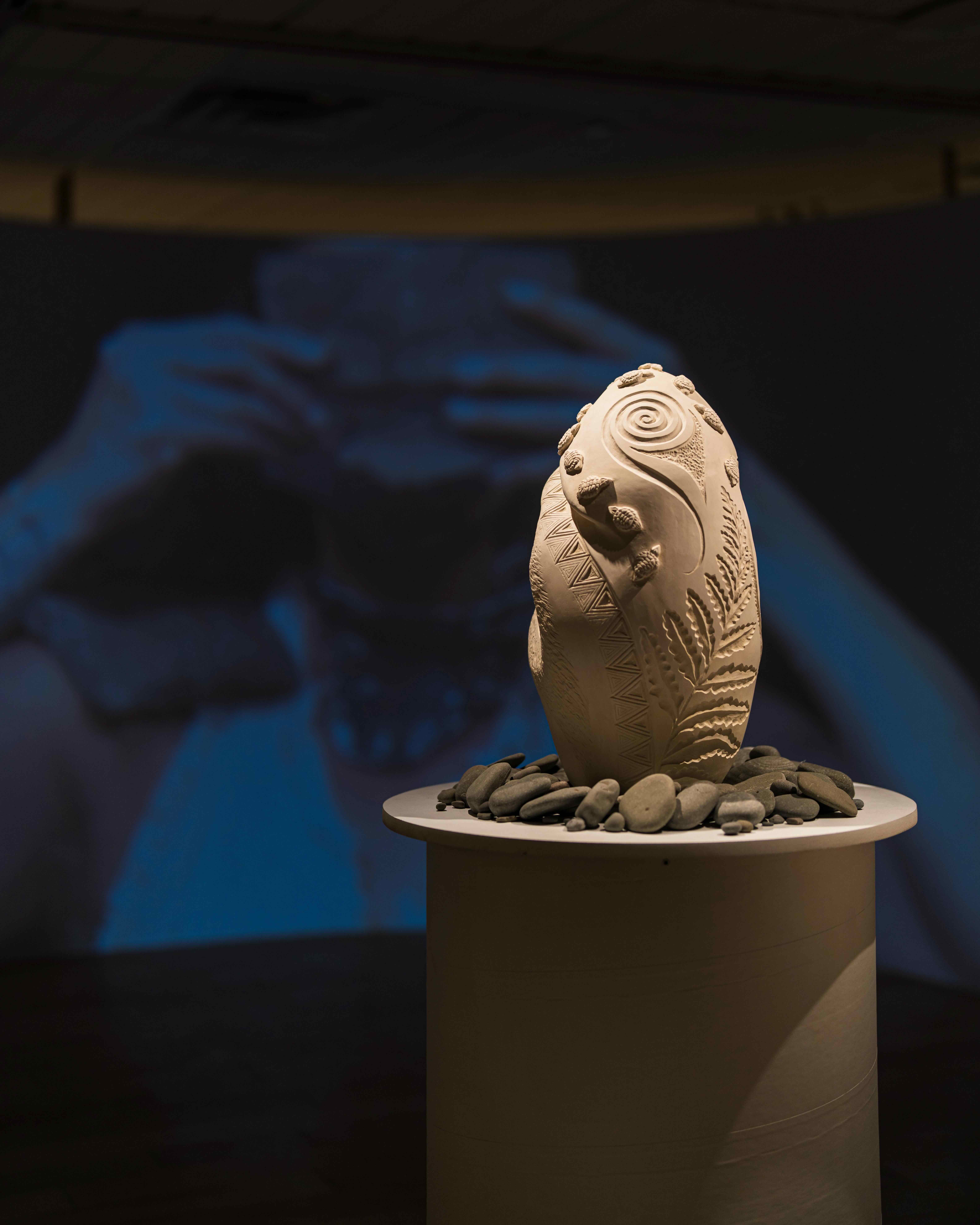 A carved ceramic sculpture with spiral and leaf-like patterns sits on a pedestal surrounded by smooth stones, with a blurred video projection in the background.